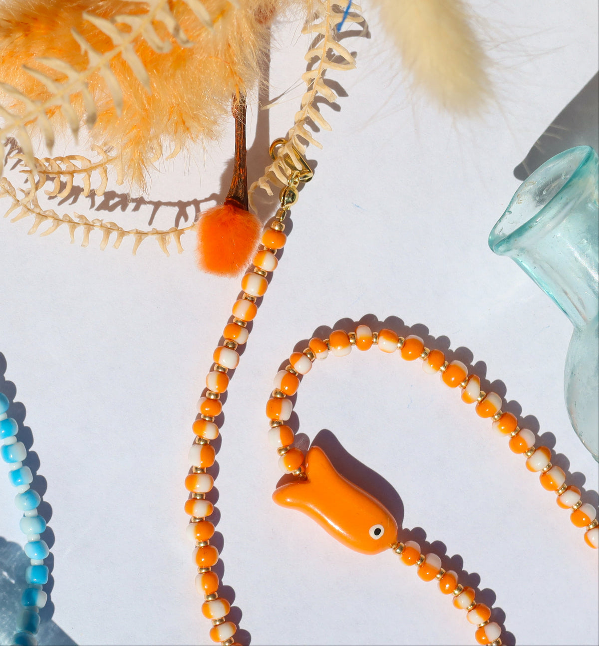 Two beaded necklaces with fish-shaped beads, one light blue and one orange, styled on a white surface with decorative dried grass, a small glass bottle, and a blue textured prop.