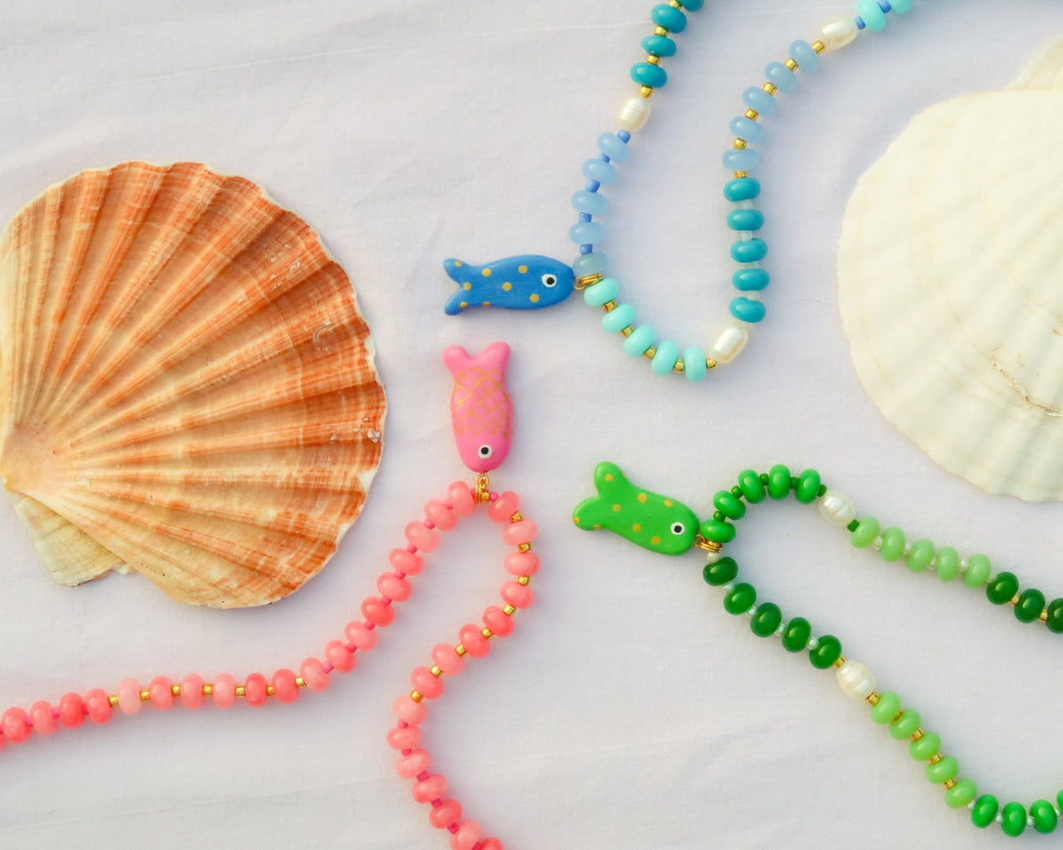 Colorful beaded necklaces with fish charms on a light background with shells.