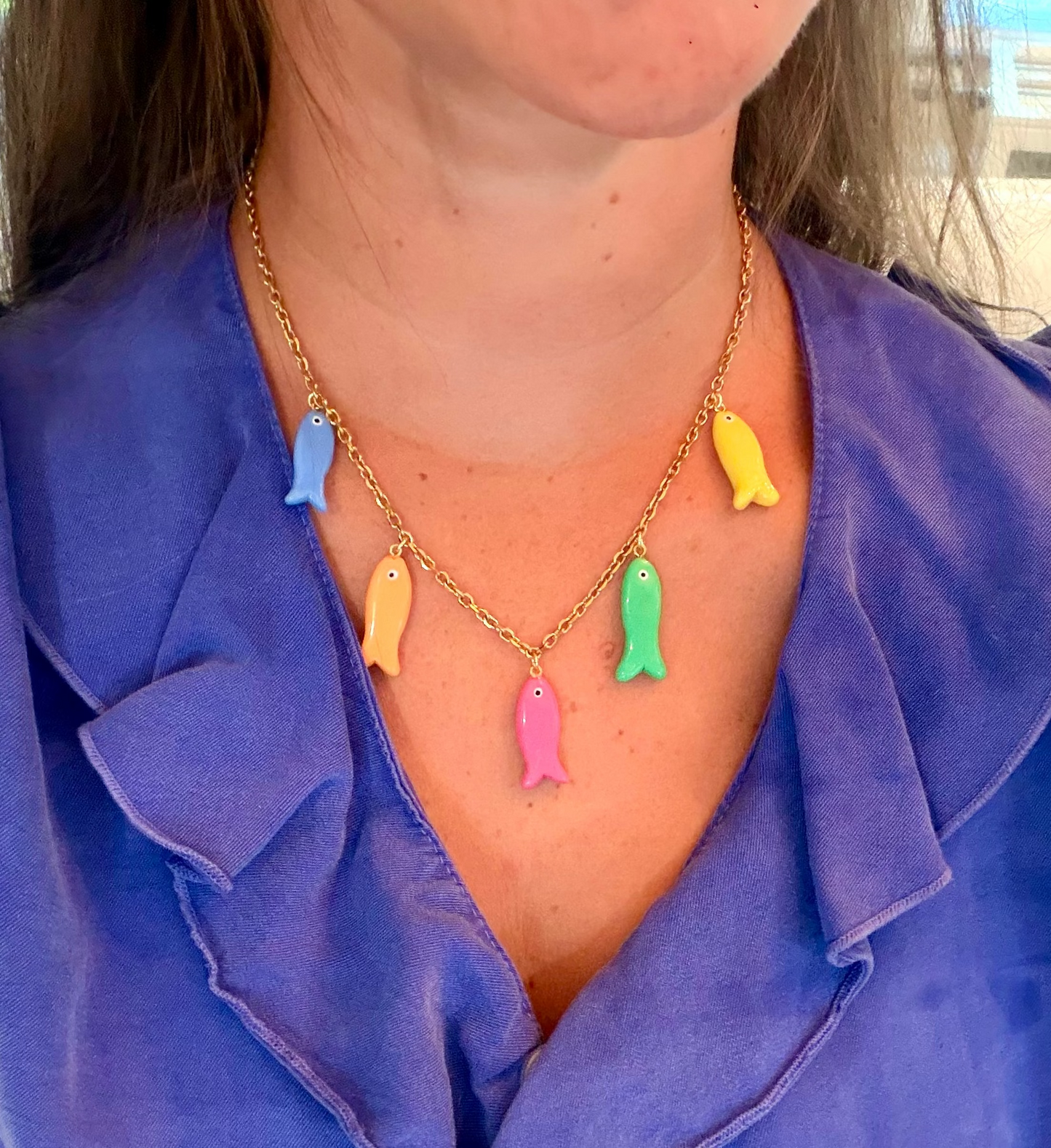A gold chain necklace with five colorful fish-shaped charms in blue, yellow, orange, green, and pink, worn by a person wearing a purple blouse.