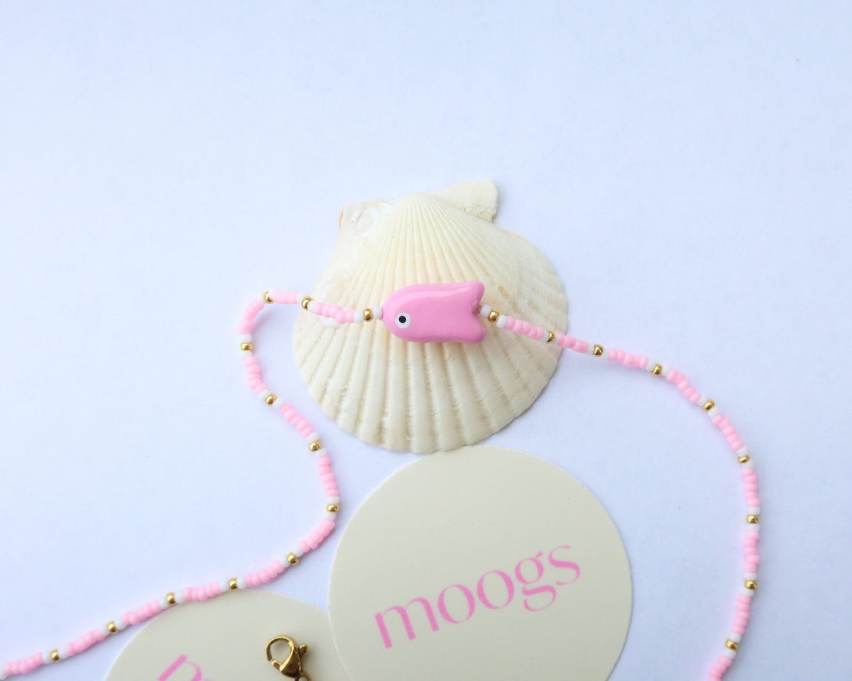 A pink beaded necklace with a small pink fish charm, displayed on a cream-colored seashell with gold spacer beads. The necklace is laid out on a white background, with part of a cream-colored circular card printed with the brand name 'moogs' visible.