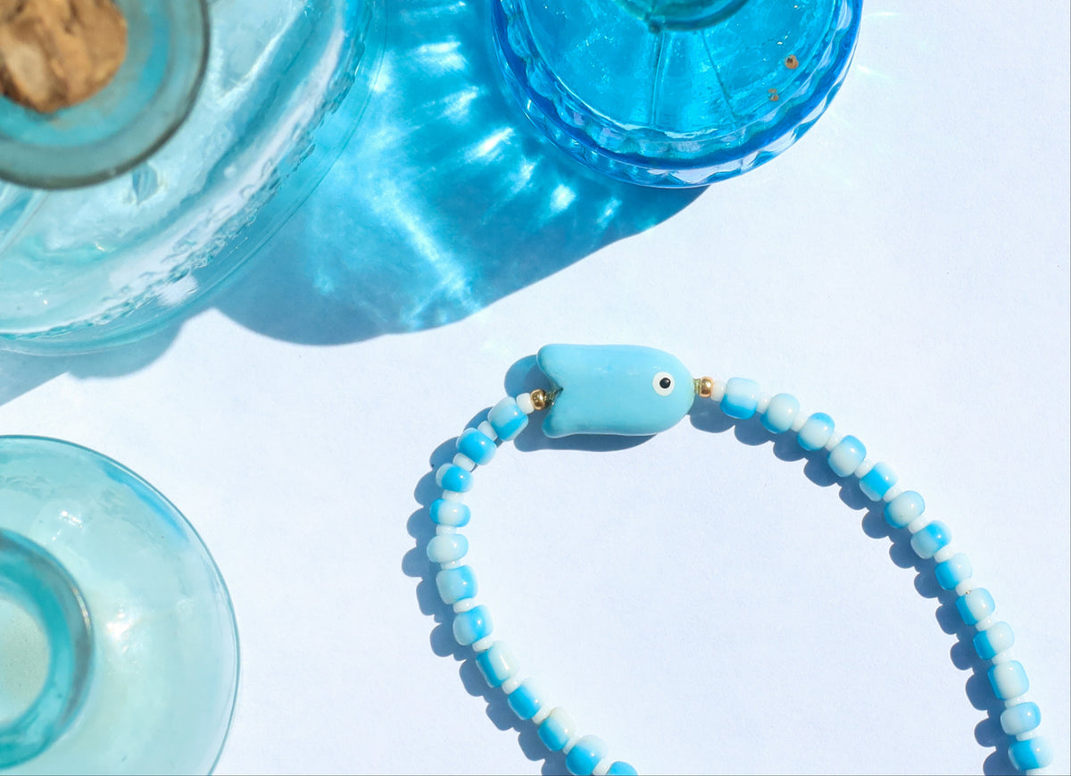 A light blue beaded bracelet with a fish-shaped charm and small gold spacer beads, arranged on a white surface next to translucent blue glass bottles.
