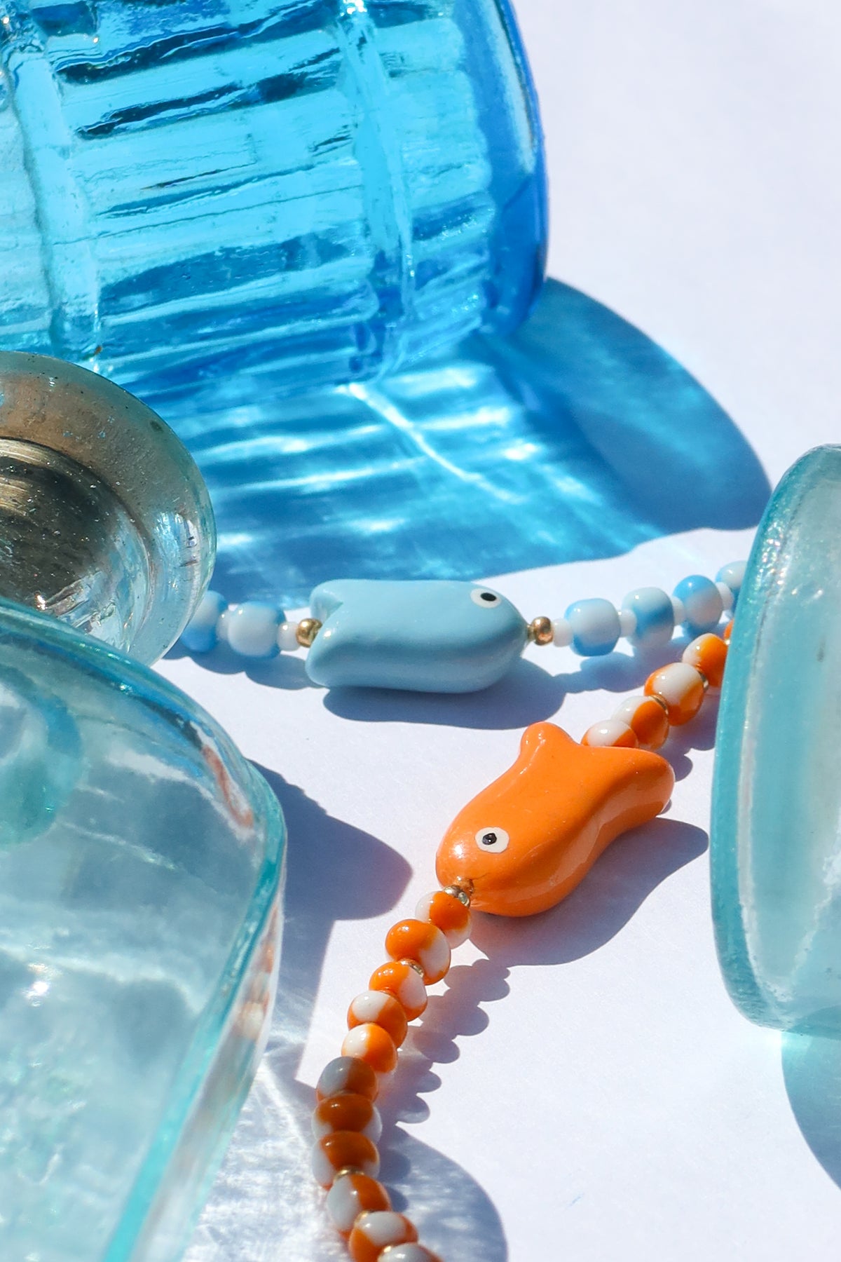 A close-up styled product shot of a beaded necklace with a light blue fish-shaped bead and orange-and-white bead strands, surrounded by translucent blue glass props on a white background.