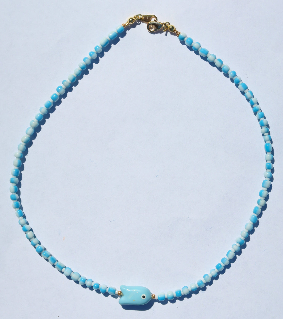 A beaded necklace with light blue and white marbled beads, featuring a small turquoise-blue fish charm with an eye, and a gold clasp, displayed on a white background.