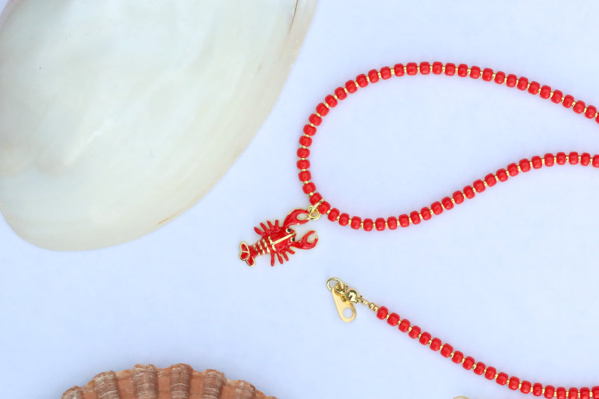 A red beaded necklace with a lobster charm and gold-tone clasp, styled on a white surface with seashell props.