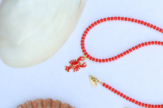 A red beaded necklace with a lobster charm and gold-tone clasp, styled on a white surface with seashell props.