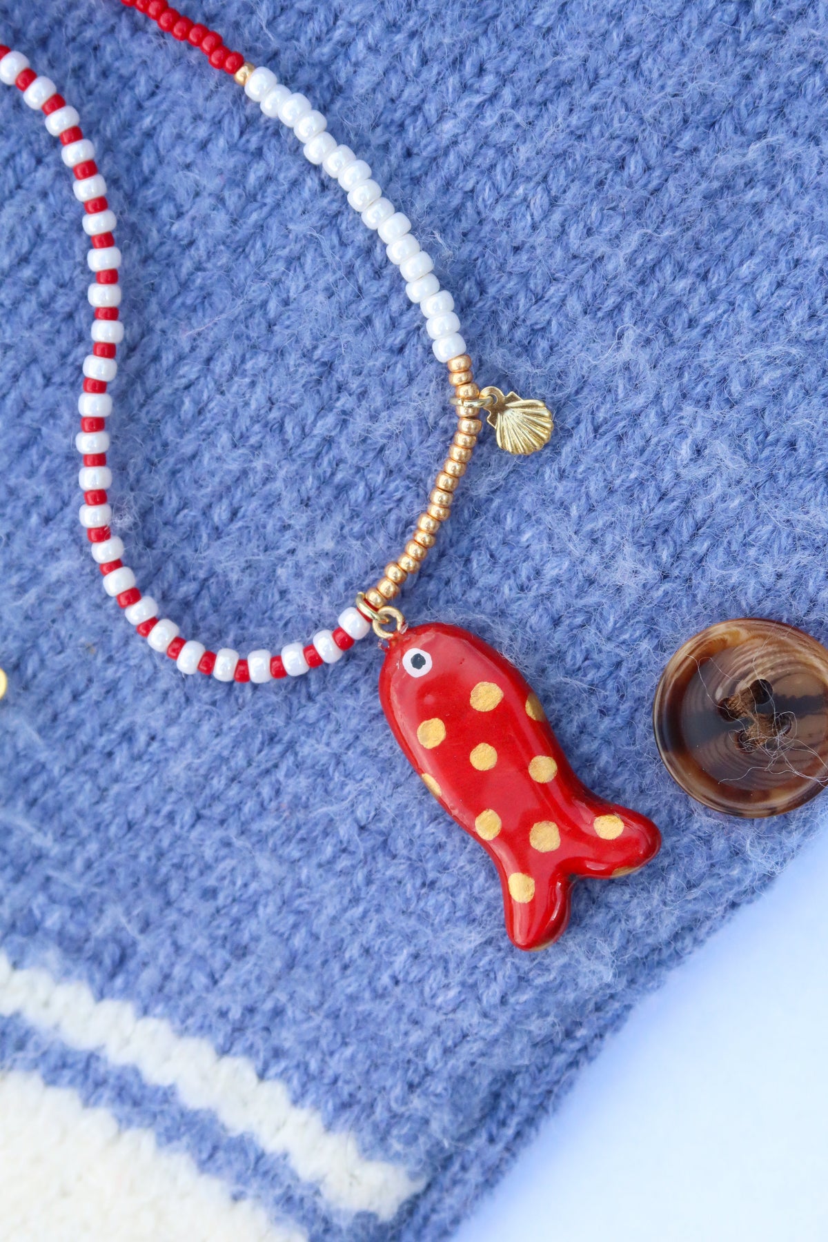 A close-up of a handmade beaded necklace with a red and white striped section, a white beaded section, gold-toned beads, a small gold shell charm, and a red fish-shaped pendant with yellow polka dots, displayed on a blue knit fabric.