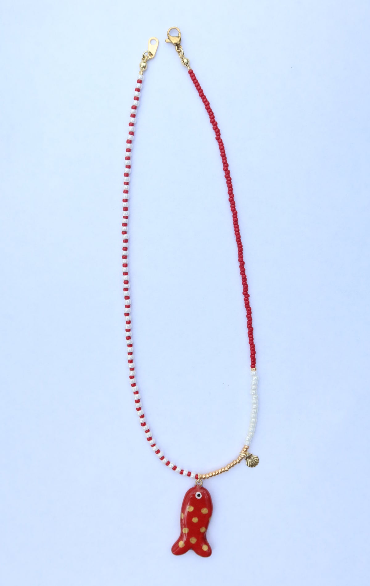 A handmade beaded necklace featuring a red fish-shaped pendant with gold polka dots, suspended from a split beaded chain of red and white beads with gold-toned hardware, set against a light blue background.